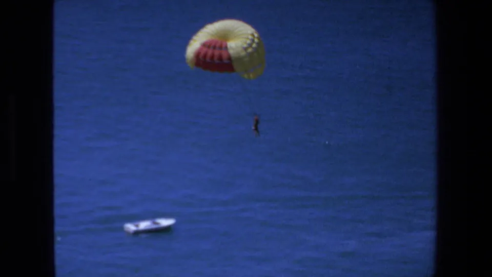 1972 Mazatlan 8mm frame showing a distant parasailer suspended beneath a red-and-yellow canopy, with a white boat lower left against the blue ocean surface.