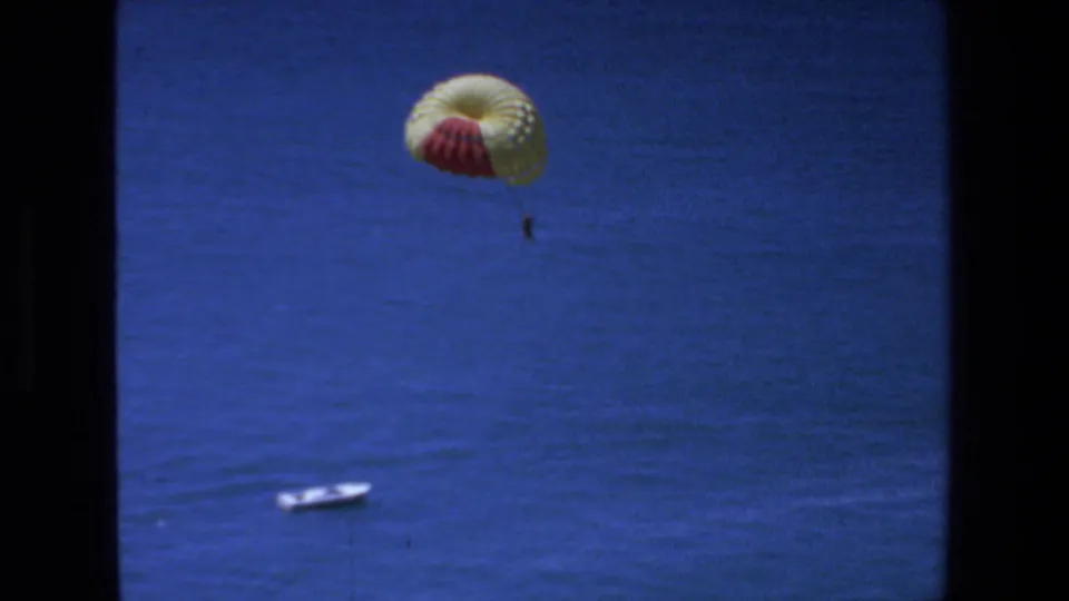 1972 Mazatlan 8mm frame with a red-and-yellow parasail canopy and a distant suspended person above open blue water, while a small white boat sits lower in the shot.
