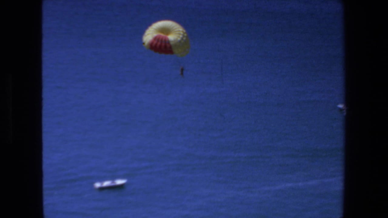 1972 Mazatlan home-movie frame showing a distant person suspended beneath a yellow-and-red parachute above deep blue ocean, with a small boat below and dark film borders at the sides.