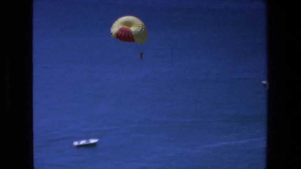 1972 Mazatlan home-movie frame showing a distant person suspended beneath a yellow-and-red parachute above deep blue ocean, with a small boat below and dark film borders at the sides.