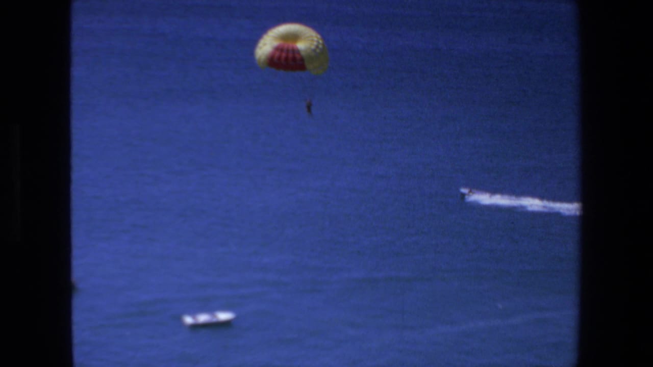 1972 Mazatlan frame with a distant parachutist above the ocean, a small white boat lower in frame, and a speedboat leaving a bright wake to the right.