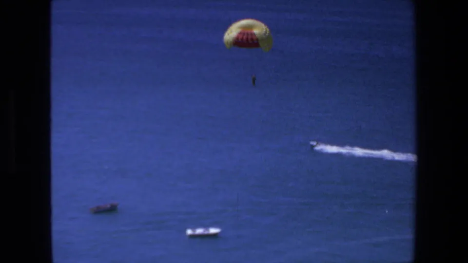 1972 Mazatlan home-movie frame showing the parachute lower over open water, with the suspended person visible beneath it, two small boats near the bottom, and a white wake crossing the right side.