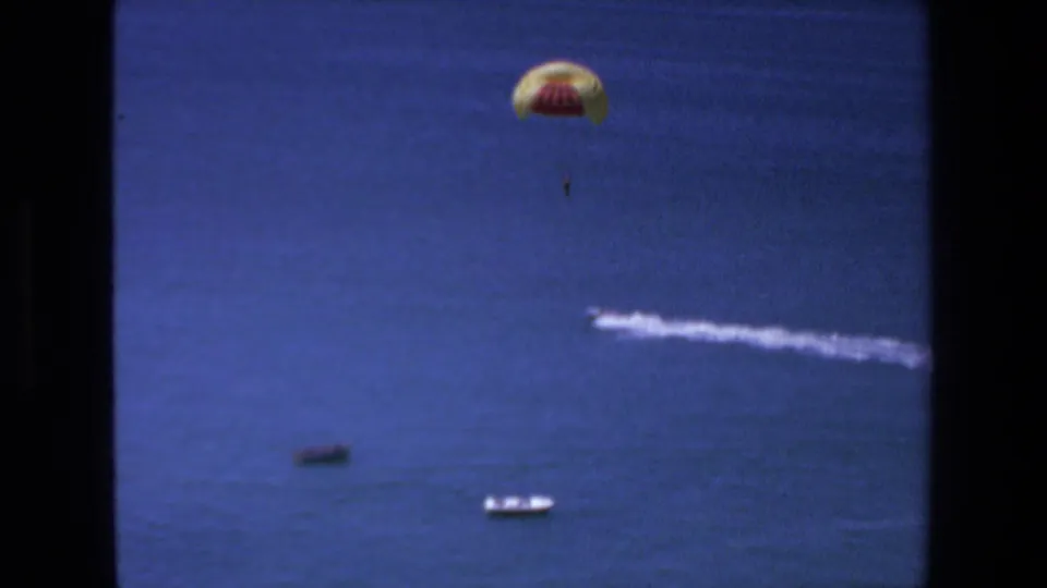 1972 Mazatlan frame showing a distant parachutist over blue ocean, with small boats near the lower edge and a long white speedboat wake stretching across the water.