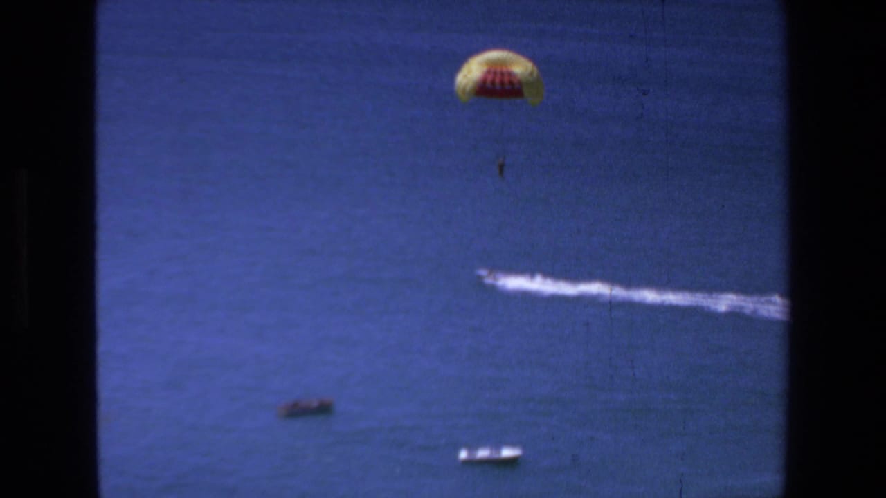 1972 Mazatlan home-movie frame showing a yellow and red parasail above deep blue water while a towing boat leaves a white wake in the distance.