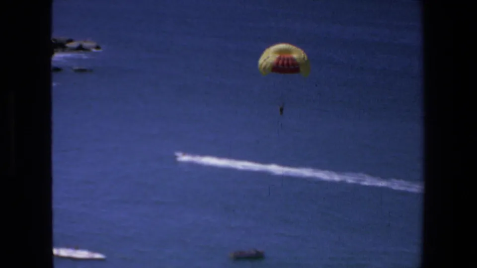 The parasail remains centered over the blue water as the towing wake passes below, with small boats and a rocky coastline visible along the left edge.