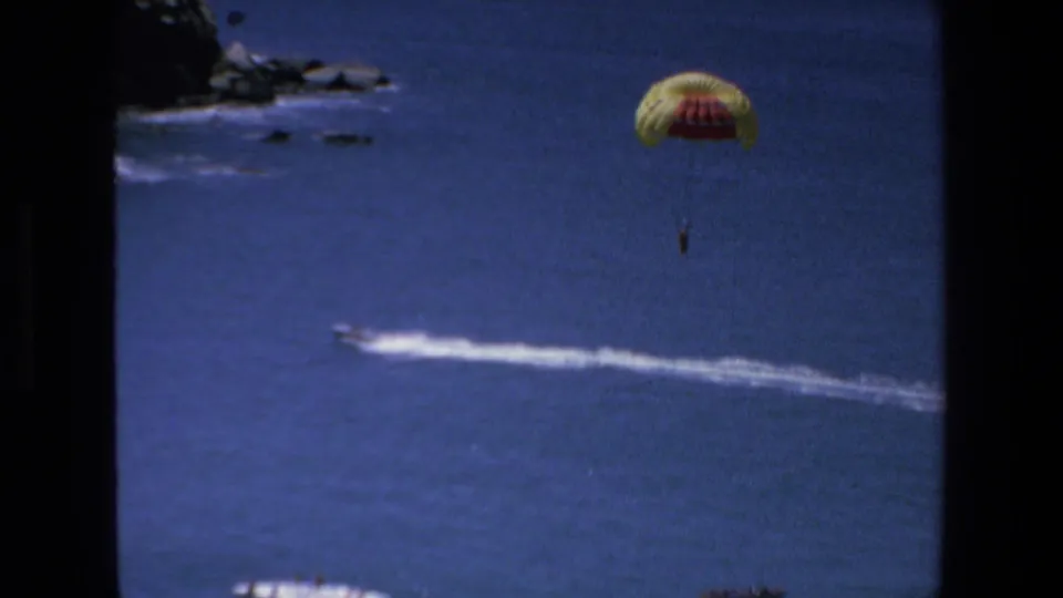 A yellow and red parasail carries a distant rider over open blue water while the towing boat's white wake curves across the lower part of the frame.