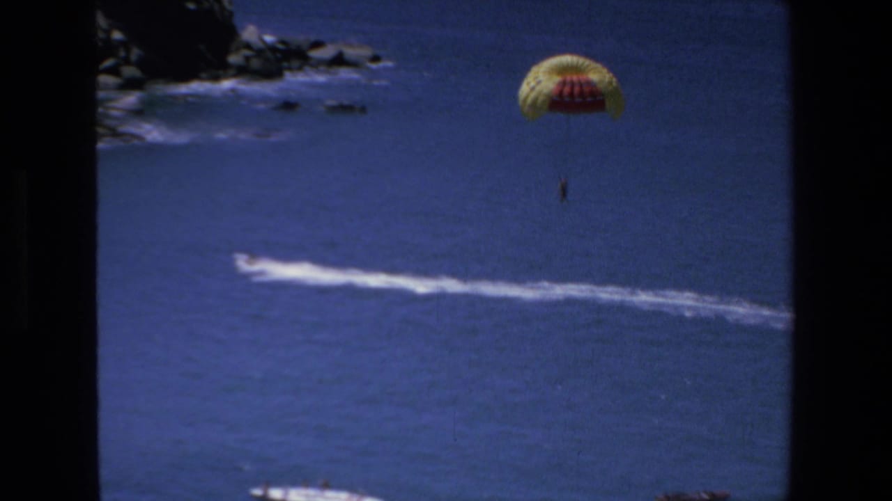 A distant person parasails under a yellow and red canopy over the blue sea in Mazatlan, Mexico, in 1972 home-movie footage, with a white tow wake below.