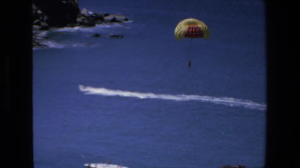 A distant person parasails under a yellow and red canopy over the blue sea in Mazatlan, Mexico, in 1972 home-movie footage, with a white tow wake below.