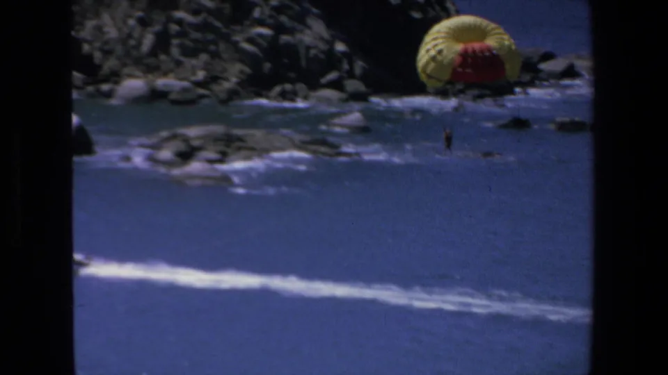 A distant parasailer appears closer to the rocky Mazatlan shoreline in 1972 8mm footage, with surf lines and deep blue water filling the frame.
