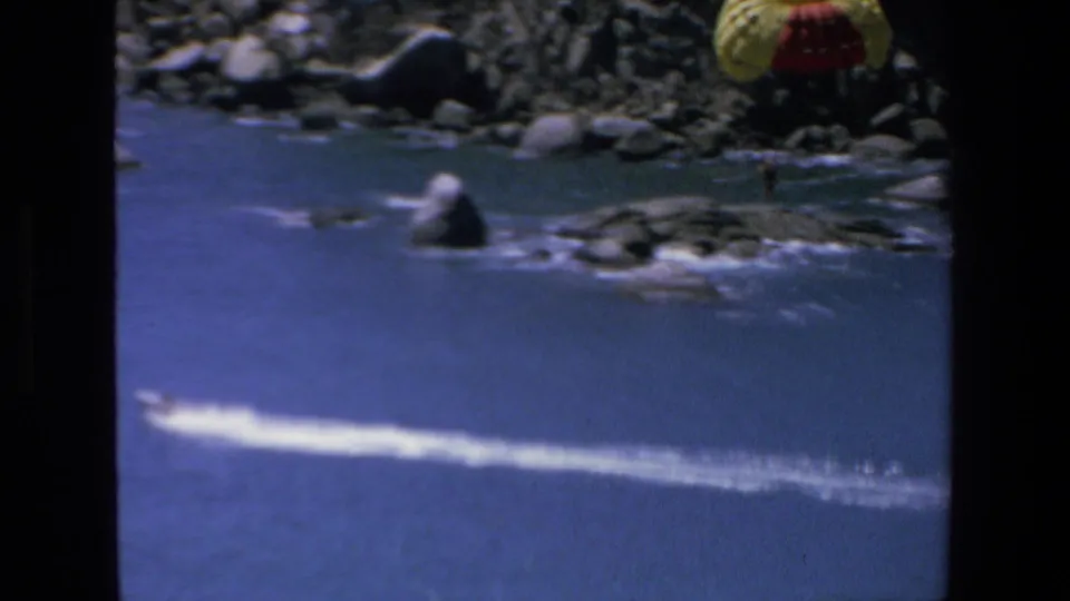 The parasail moves partly out of frame above a rocky Mazatlan shoreline, while blue water, white surf, and the tow wake remain visible in the 1972 home movie.