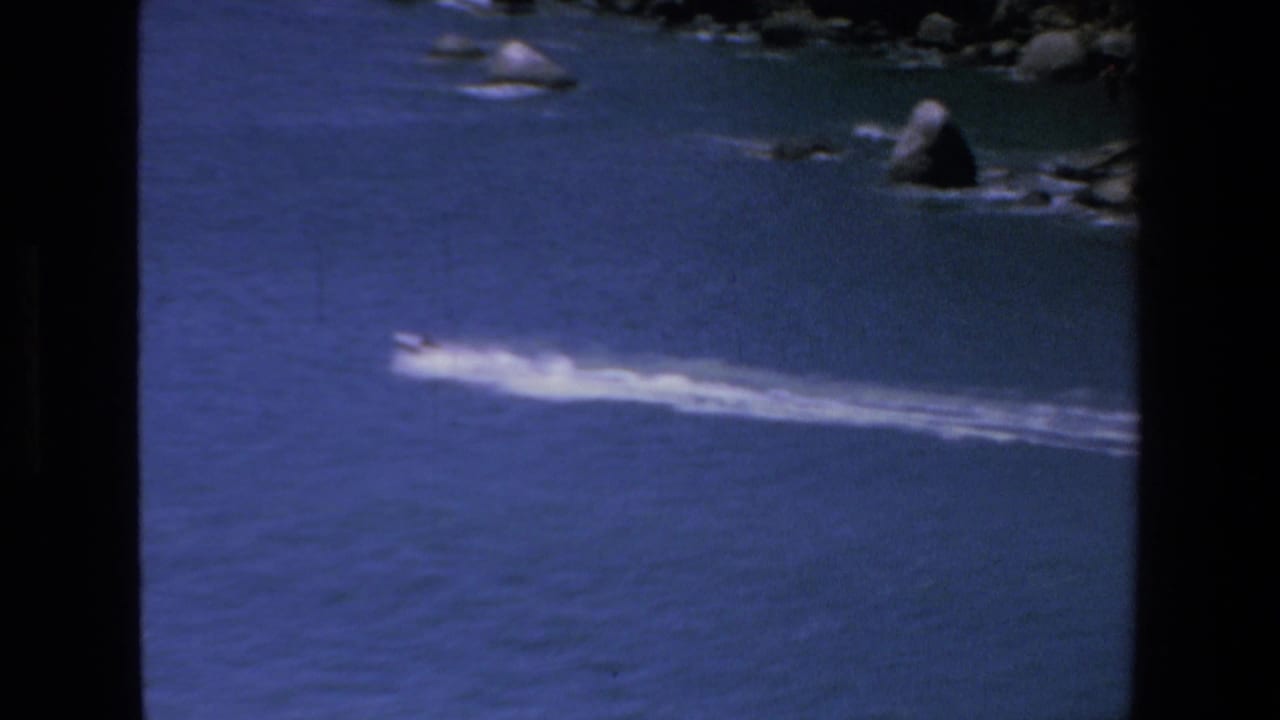 In Mazatlan, Mexico in 1972, a small speedboat moves across deep blue water, leaving a long white wake below a rocky shoreline.