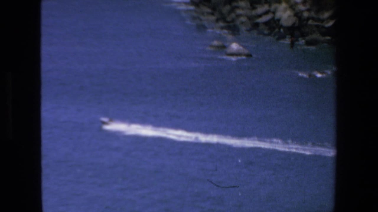 A distant speedboat cuts across blue water in the Mazatlan 1972 home-movie frame, with a pale wake trailing behind and shoreline rocks at the top.