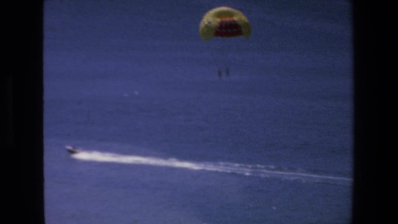 1972 Mazatlan home-movie frame showing a yellow and red parasail high over blue water while a motorboat speeds across the ocean below.