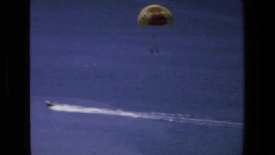1972 Mazatlan home-movie frame showing a yellow and red parasail high over blue water while a motorboat speeds across the ocean below.