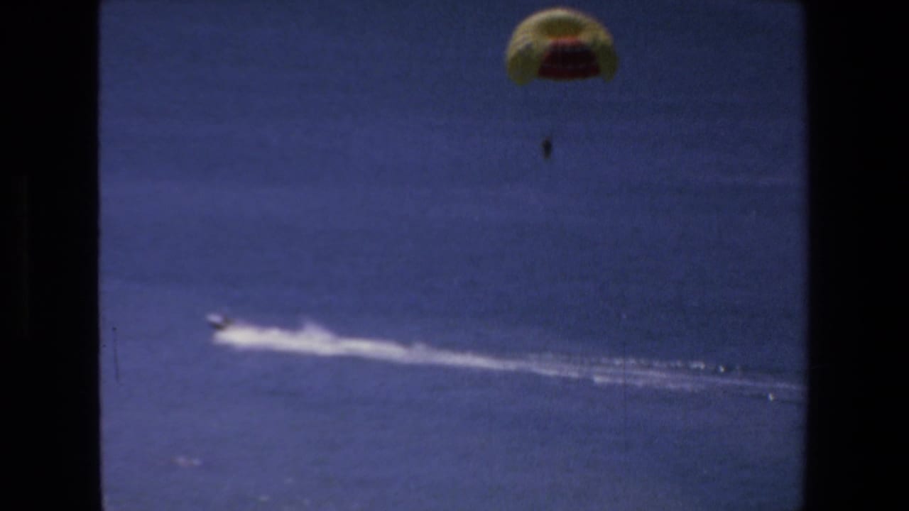 1972 Mazatlan 8mm frame with a distant parasailer above the ocean and a small motorboat pulling a long white wake across the water.