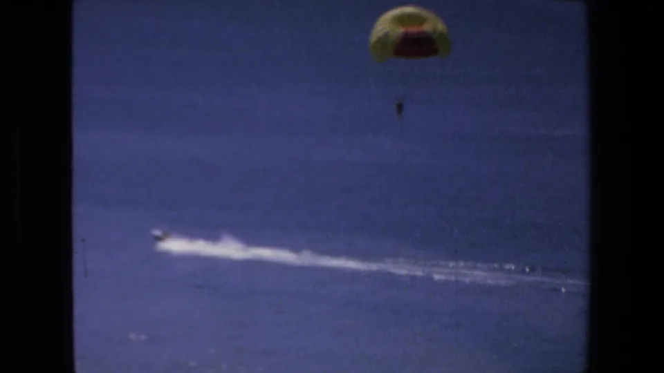 1972 Mazatlan 8mm frame with a distant parasailer above the ocean and a small motorboat pulling a long white wake across the water.