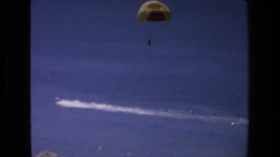 Distant 1972 Mazatlan vacation footage frame showing a parasail and rider above open water while the towing boat cuts a white path below.