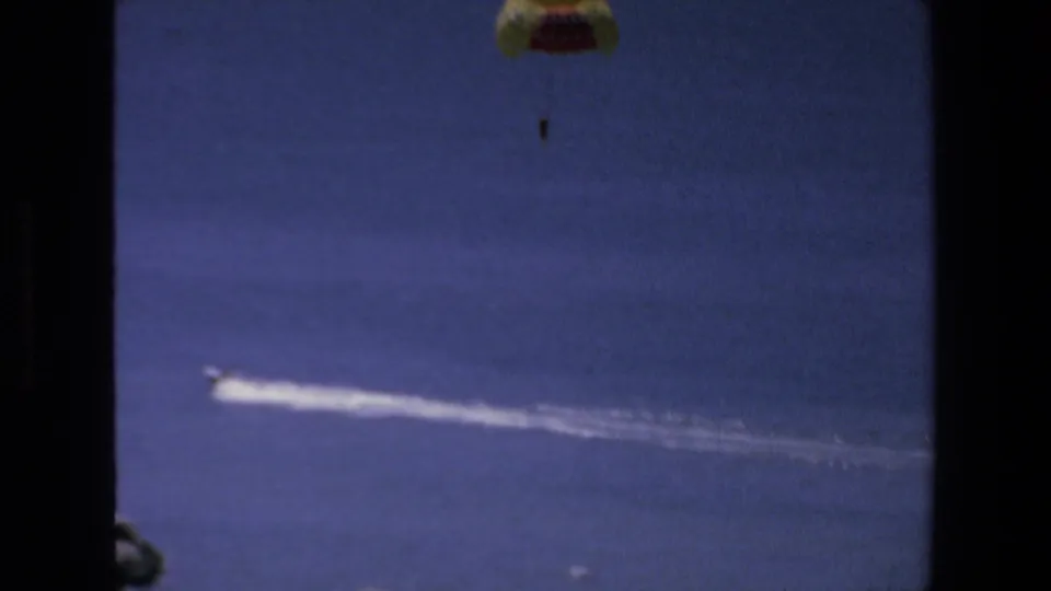 Final 1972 Mazatlan 8mm frame with the parasail near the top edge, a distant rider below it, and a motorboat wake running across the blue ocean.