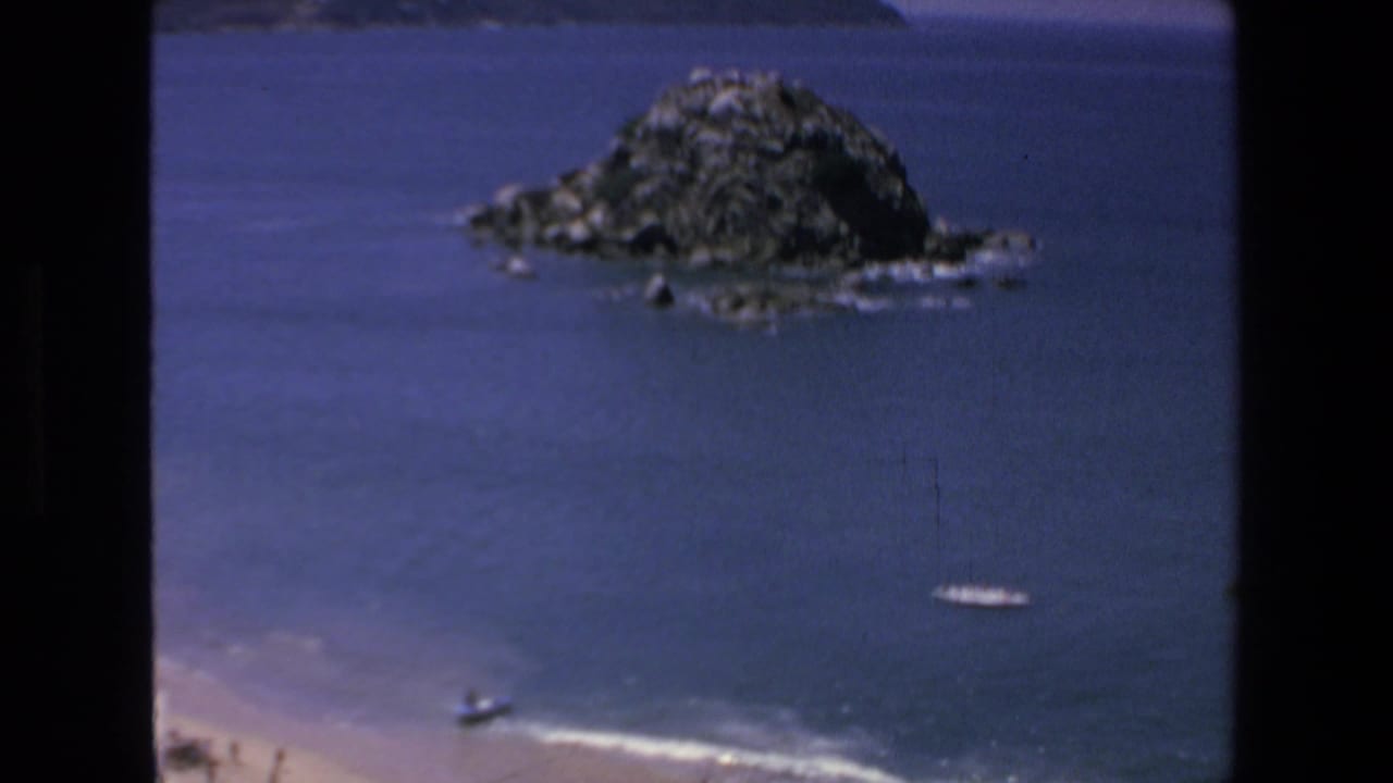 1972 Mazatlan home-movie frame showing a rocky island offshore, blue ocean, surf near the beach, and small boats close to the shoreline.