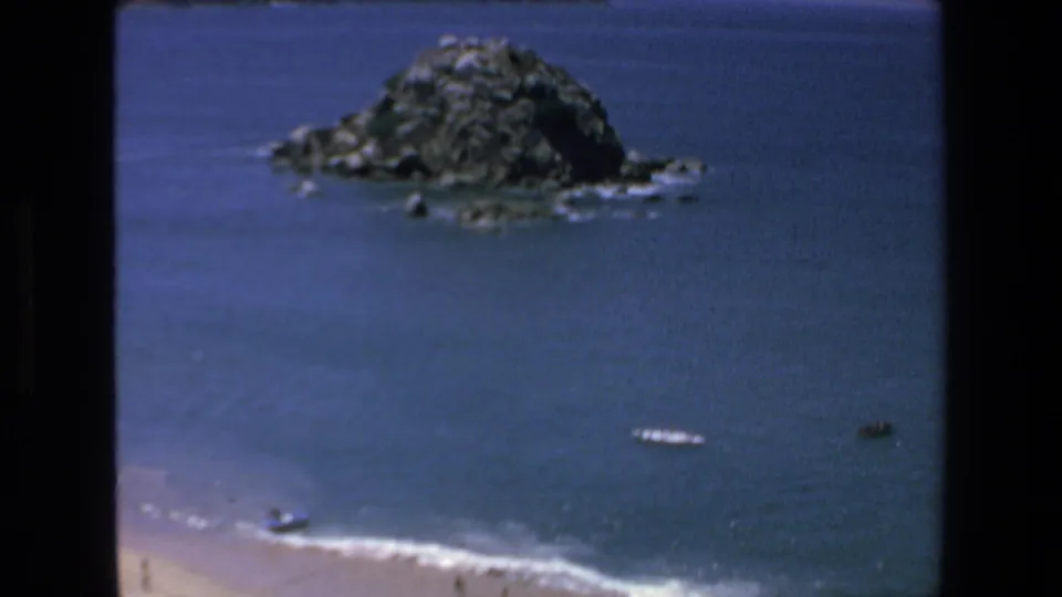 1972 Mazatlan coastal view with a rocky island rising from the ocean and small boats scattered near the beach and surf line.