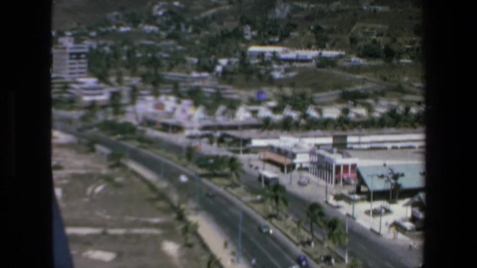Elevated 1972 Mazatlan home-movie view over a coastal road with cars, low-rise buildings, palm trees, open lots, and hills in the background.