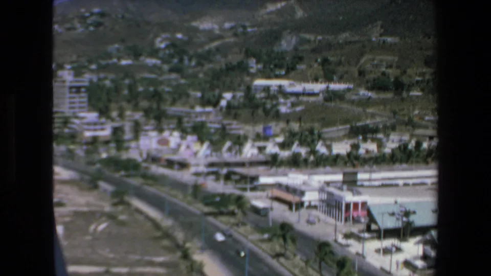 1972 Mazatlan frame looking down on a busy coastal highway, nearby buildings, palms, hillside vegetation, and scattered traffic in a seaside town setting.