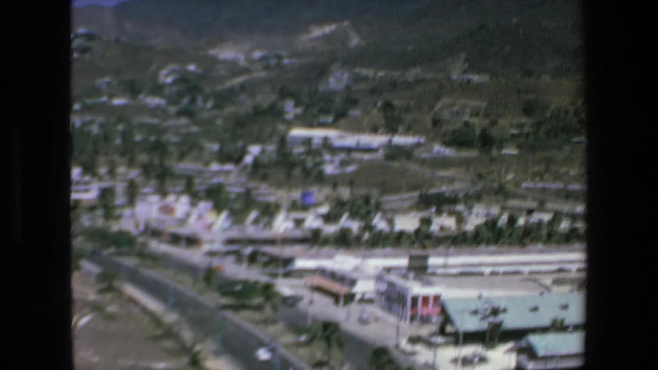 1972 Mazatlan aerial 8mm frame showing a large green-roofed hotel complex near a road, with palms, low buildings, and green hills beyond.