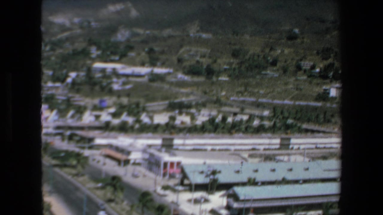 1972 Mazatlan aerial frame with a modern hotel roof in the foreground, roadway access, palm landscaping, and scattered homes on the hillside behind.