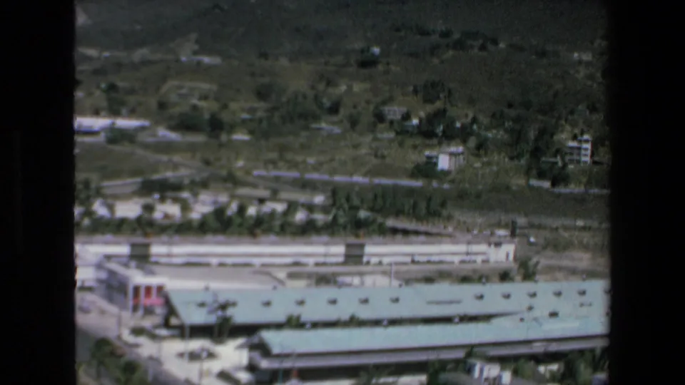 1972 Mazatlan 8mm aerial frame showing a long green-roofed hotel or commercial building, nearby roadway, palms, and residential structures on the green hillside.