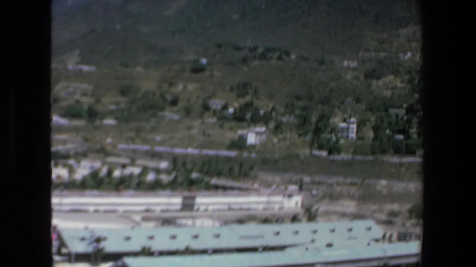 1972 Mazatlan aerial 8mm frame looking across a road and green-roofed complex toward scattered hillside buildings and open vegetation.