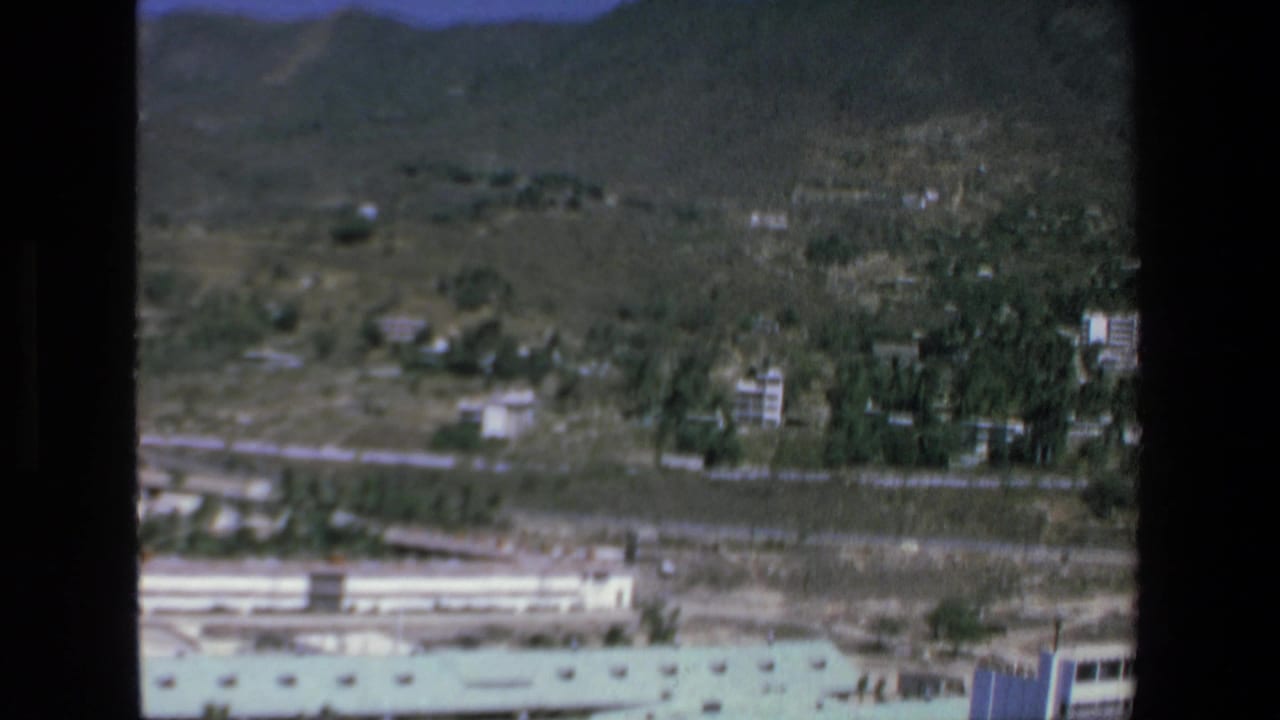 1972 Mazatlan 8mm aerial frame showing an arid hillside landscape with scattered low-rise buildings, green vegetation, distant mountains, and a light-colored roofline at the bottom edge.