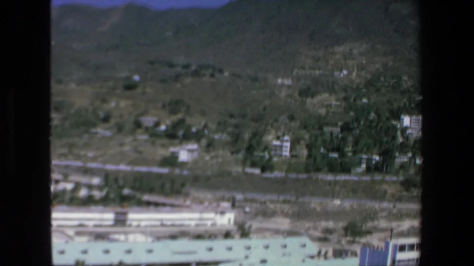 1972 Mazatlan 8mm aerial frame showing an arid hillside landscape with scattered low-rise buildings, green vegetation, distant mountains, and a light-colored roofline at the bottom edge.