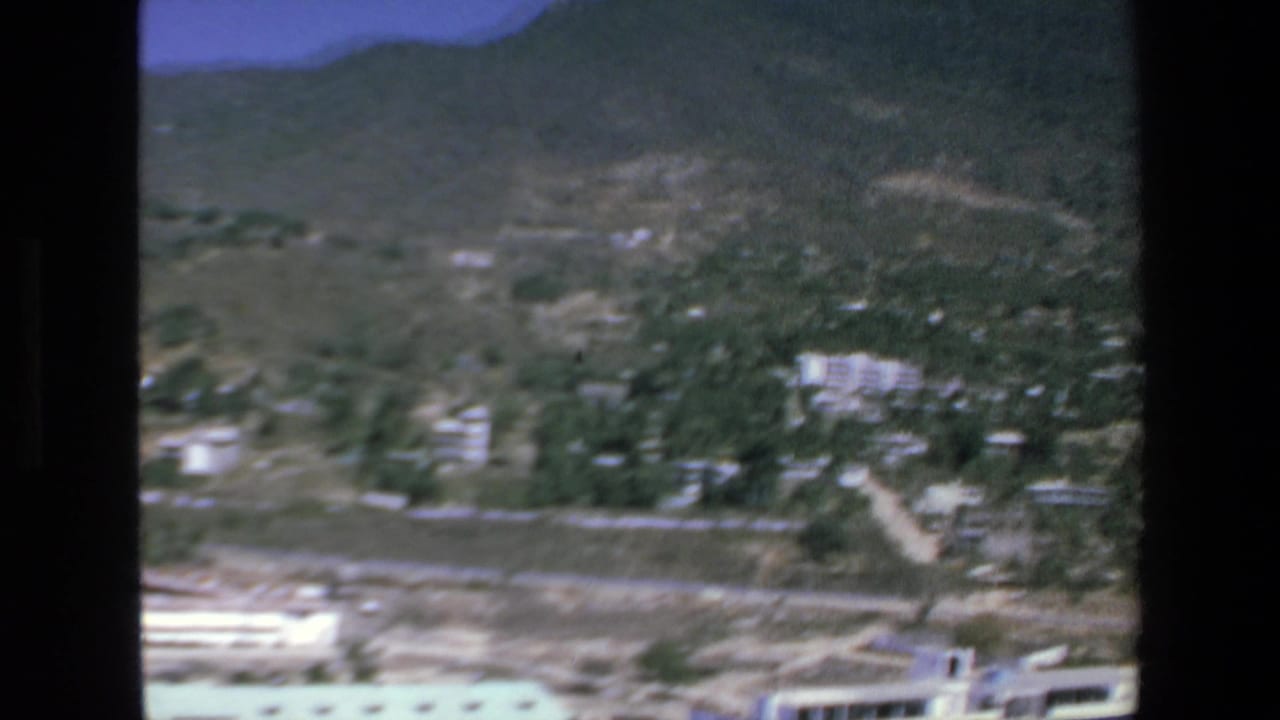 1972 Mazatlan aerial frame with hillside terrain, scattered structures among vegetation, dry open land, and a bright low building near the lower frame edge.
