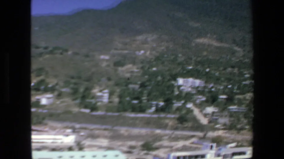 1972 Mazatlan aerial frame with hillside terrain, scattered structures among vegetation, dry open land, and a bright low building near the lower frame edge.