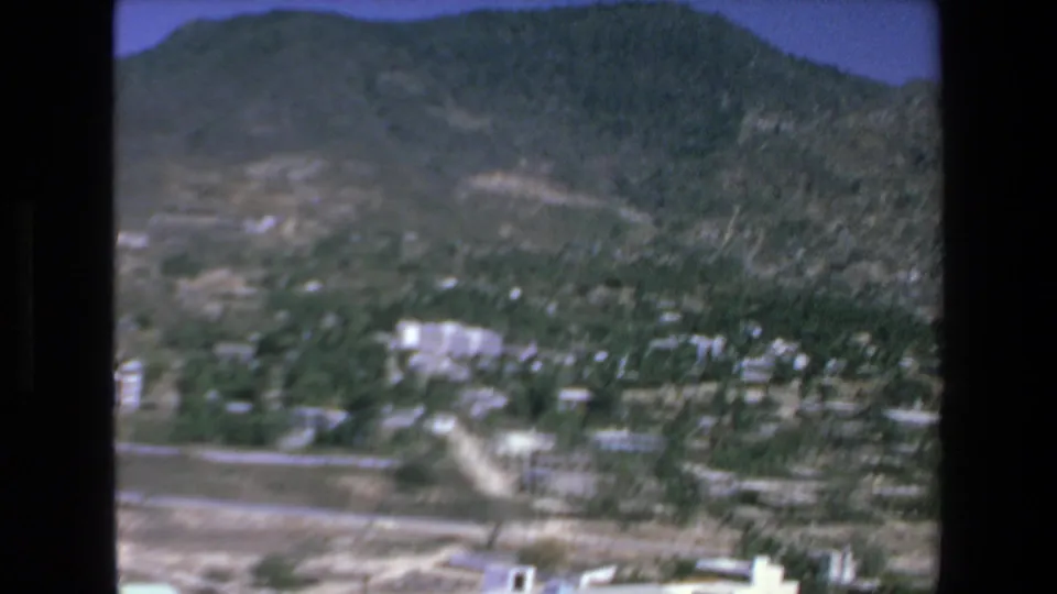 Soft 8mm aerial frame from Mazatlan in 1972 showing low-rise buildings, a pale road or track, green vegetation, dry ground, and mountain slopes beyond.