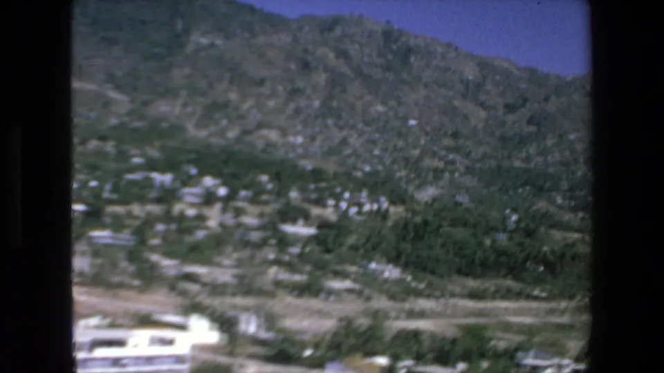 1972 Mazatlan 8mm aerial frame looking across scattered hillside buildings, green trees, dry open terrain, and rugged mountain slopes under a blue sky.
