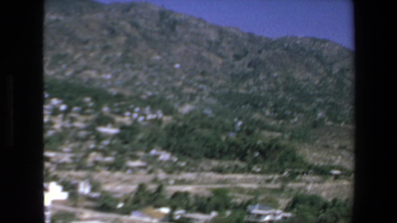 1972 Mazatlan home-movie frame looking from a mountaintop residence across a green valley, scattered buildings, and rugged hills under a blue sky.