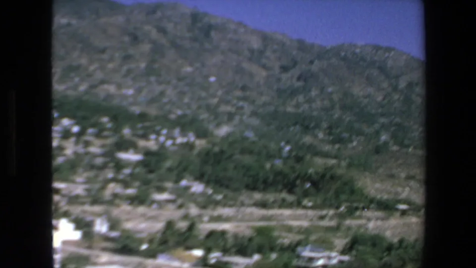 1972 Mazatlan home-movie frame looking from a mountaintop residence across a green valley, scattered buildings, and rugged hills under a blue sky.