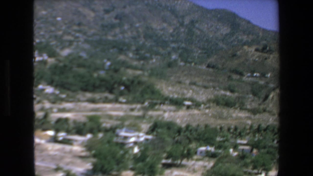 1972 Mazatlan frame showing a hillside valley with low buildings, trees, pale dirt roads, and rocky mountains in the background.