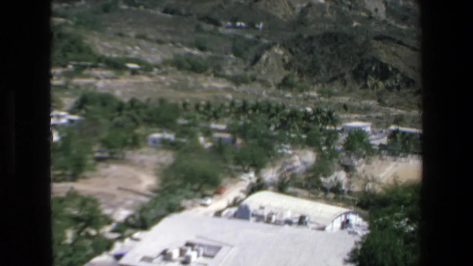 1972 Mazatlan home-movie frame panning downward toward trees, pale roads, hillside terrain, and a flat-roofed building at the lower edge of the view.