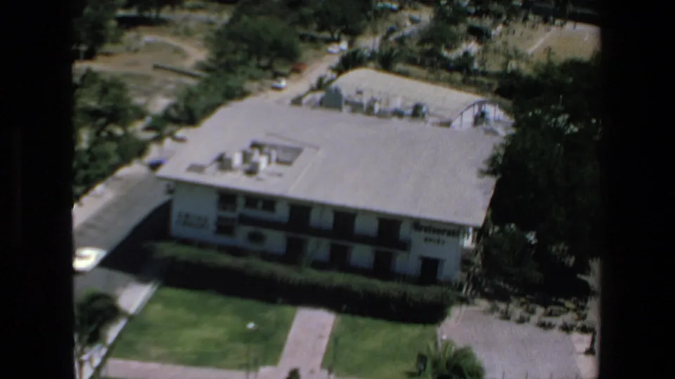 1972 Mazatlan frame showing a low flat-roofed building marked as a restaurant, a green lawn, surrounding trees, and hillside roads beyond.