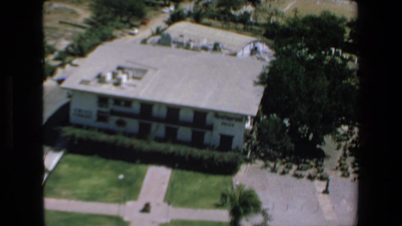 1972 Mazatlan home-movie frame showing an overhead view of a white low-rise hotel, flat roof, green lawn, hedges, and palm trees in bright sun.