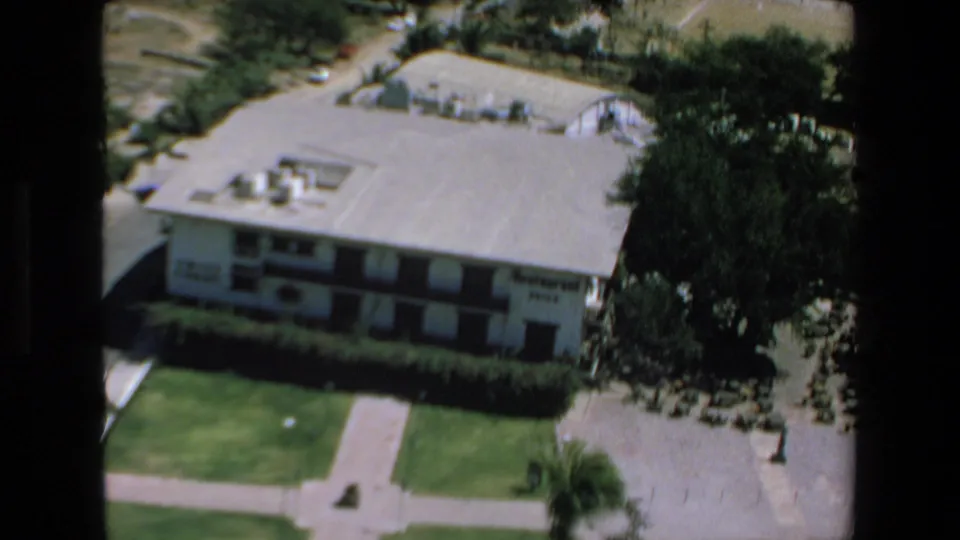 1972 Mazatlan home-movie frame showing an overhead view of a white low-rise hotel, flat roof, green lawn, hedges, and palm trees in bright sun.