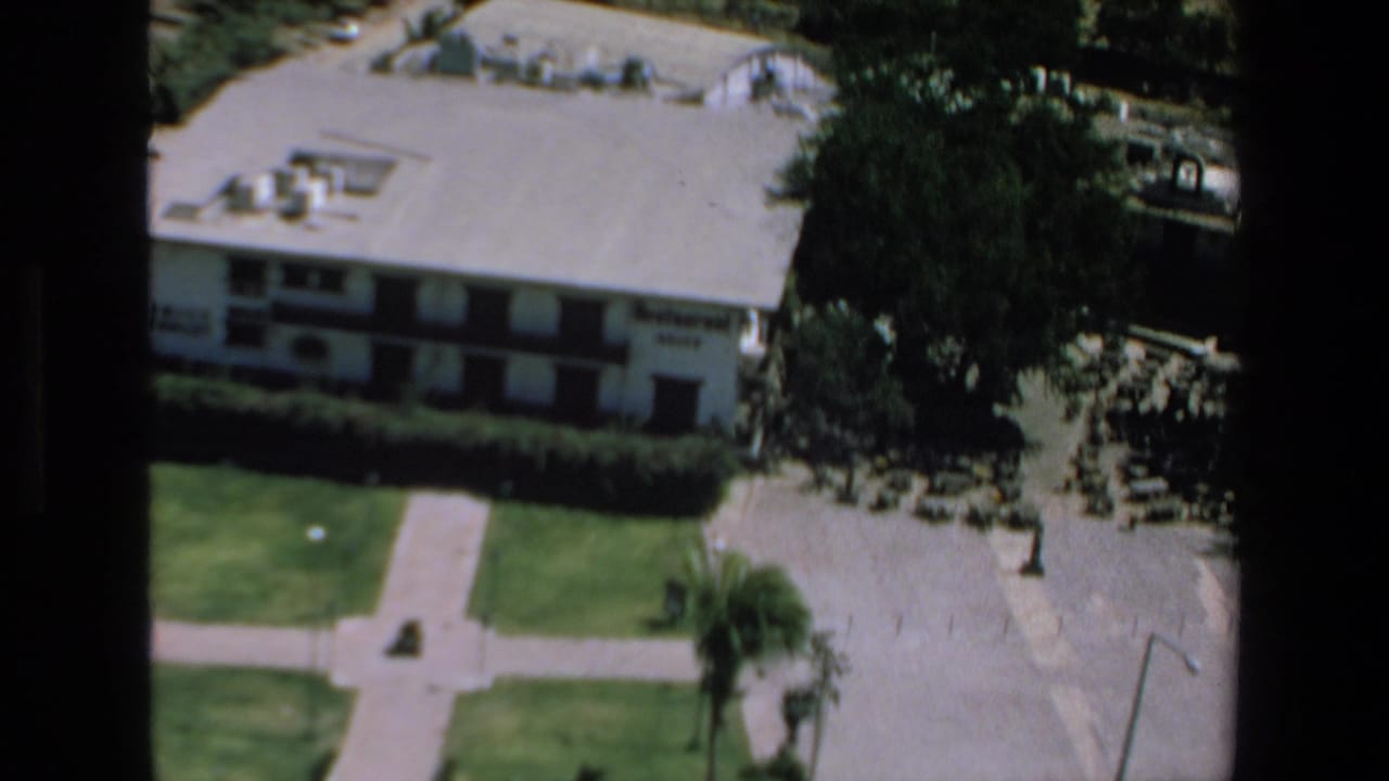 1972 Mazatlan frame with a white hotel seen from above, showing rooftop fixtures, shaded trees, lawn walkways, and adjacent pavement.