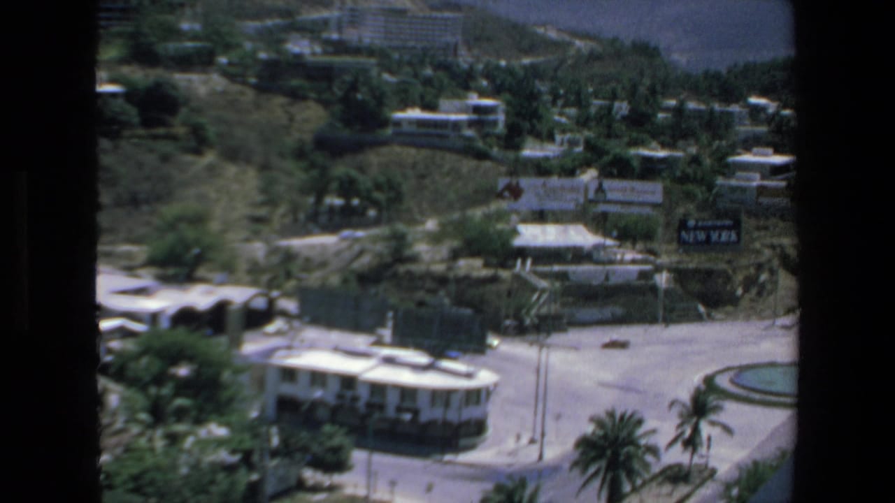 Elevated view across a hillside city area with roads, buildings, palms, cars, and a circular fountain.