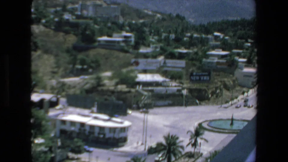 1972 Mazatlan home-movie frame with a wider balcony view across roads, small cars, hotel-like buildings, palms, hillside greenery, and a circular fountain.