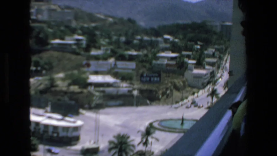 1972 Mazatlan balcony-view frame showing streets, hillside buildings, palms, a circular fountain, and a partially visible person wearing sunglasses at the right edge.