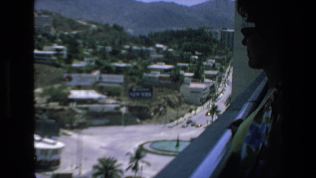 In 1972 Mazatlan home-movie footage, a woman in sunglasses stands in shadow beside a window or balcony overlooking roads, palm trees, low buildings, and distant mountains.