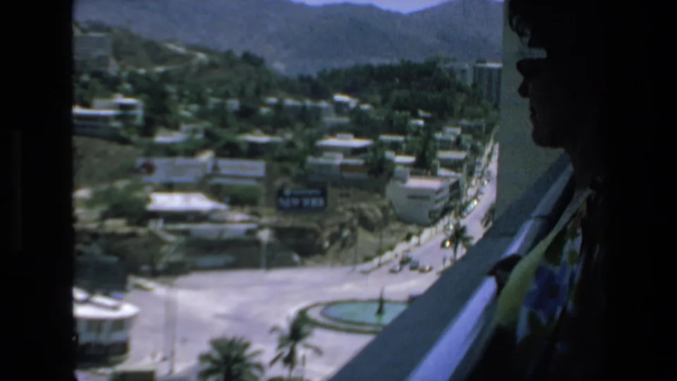 In 1972 Mazatlan home-movie footage, a woman in sunglasses stands in shadow beside a window or balcony overlooking roads, palm trees, low buildings, and distant mountains.