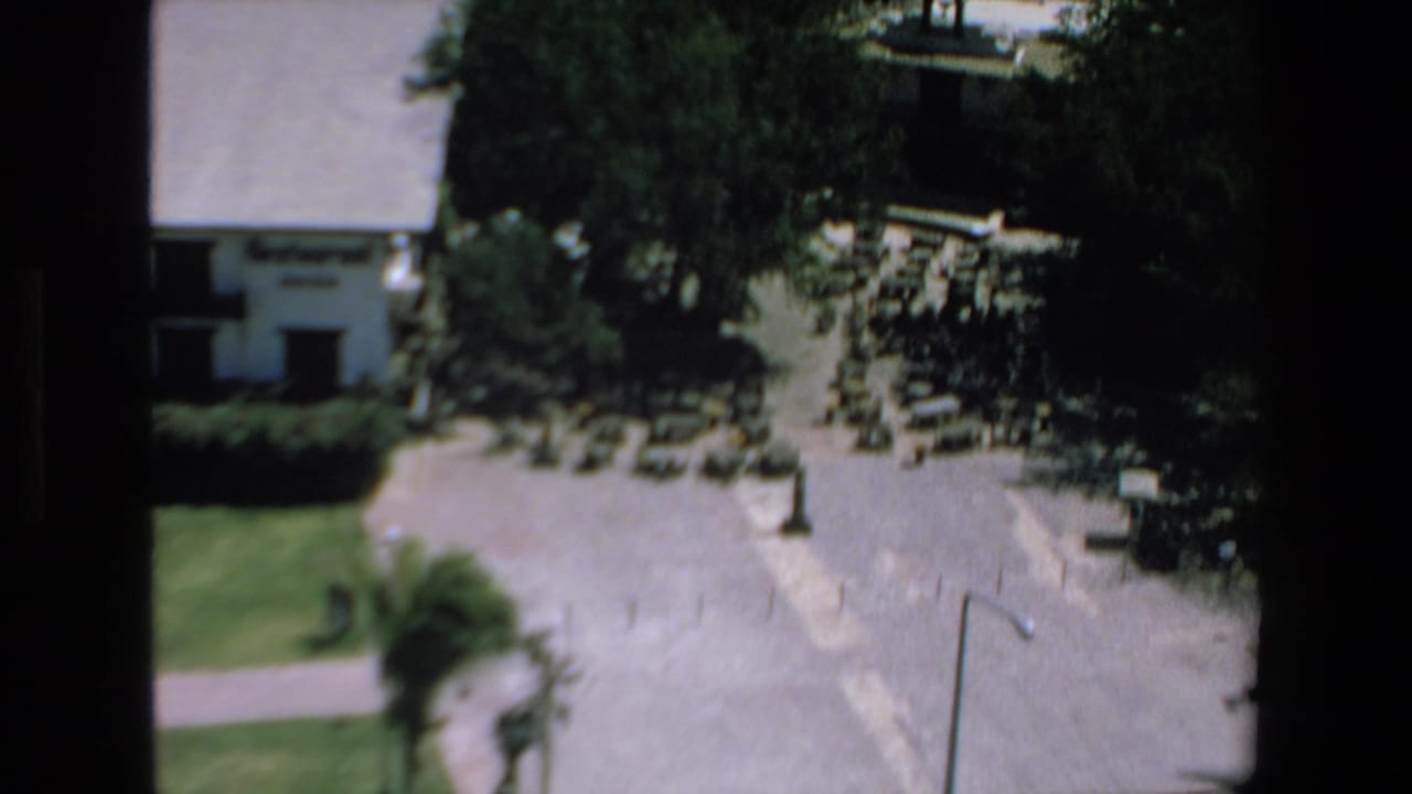 Elevated 1972 Mazatlan home-movie view of a sunlit parking area with parked vehicles, trees, and a light-colored building partially framed by 8mm film borders.